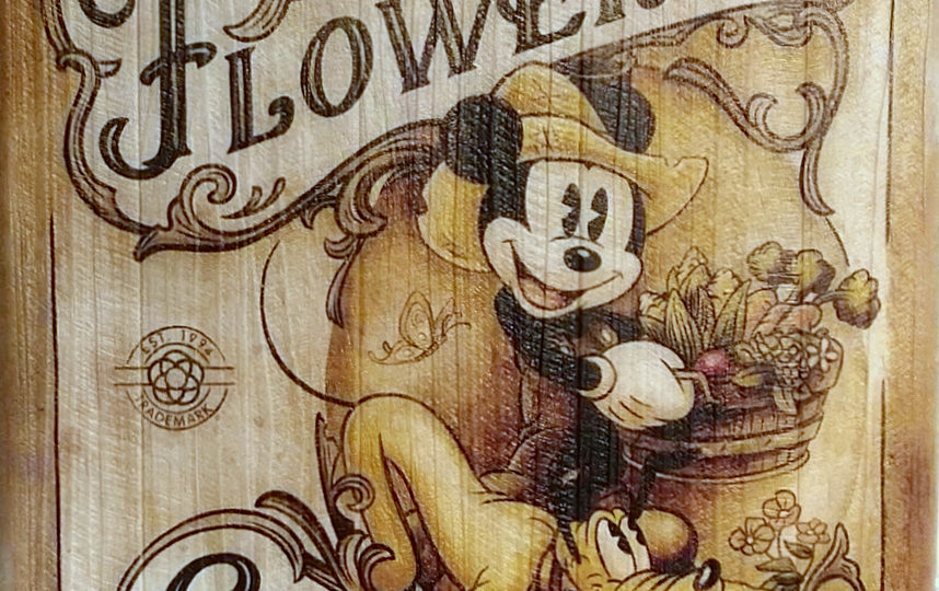 A white washed wooden sign for the Epcot International Flower and Garden Festival showing Mickey Mouse in overalls and a farm hat carrying what appears to be a buschell of corn tulips and asperagas while pluto eagerly looks on under foot.